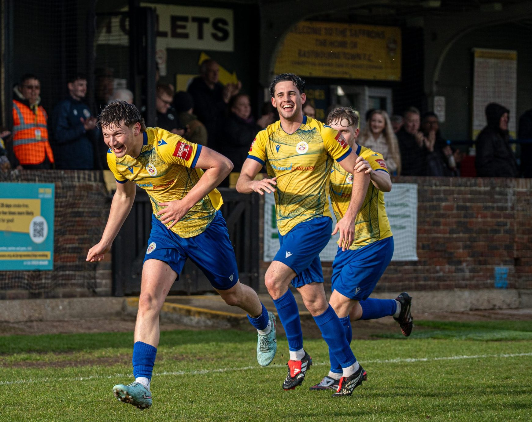 Jimmy Holman celebrates in celebration along with Sonny Walsh and Hervey Greig after Holman scores Eastbourne Town's third goal against Sheppey United at The Saffrons